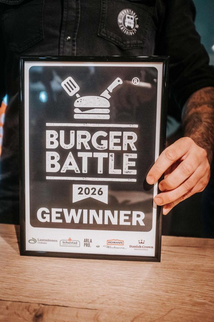Alt-Text: Eine Person haelt ein Schild mit der Aufschrift „Burger Battle 2018 Gewinner“.